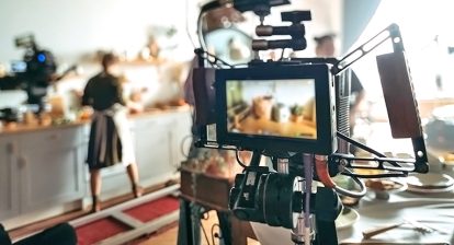 behind the scenes production of a tv show. A camera close up on some food, with a woman standing at a counter, to the left of the close up, which is out of focus.