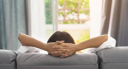 Viewed from behind, a woman relaxes on a couch and supports her head in her hands