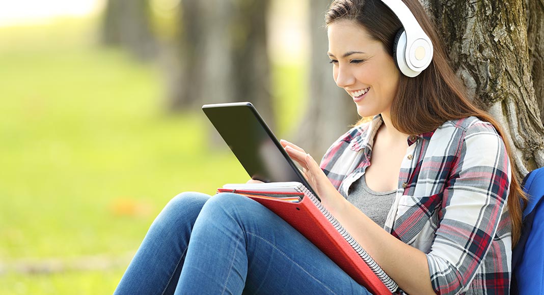 Girl sits by a tree with headphones looking at a tablet