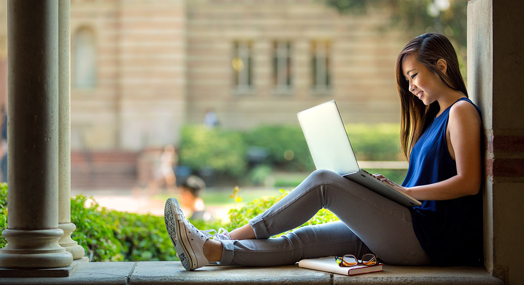 A young woman, presumably listening to her elearning course, sits outside in a school courtyard with her laptop on her lap.