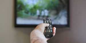 a woman's hand holding a TV remote towards the TV with a national geographic style show playing on the TV, signifying the different accents and languages found in american media and marketing