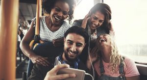 A group of four young adults gather together to listen to and watch what is on their friend's cellphone. They're all laughing and looking like they're having a good time.