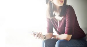 A woman sits with her phone in one hand and headphones in her ears. She appears to be listening intently.