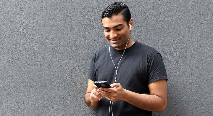 A man with dark hair and ear buds in his ears, holds his cellphone in both hands and smiles, enjoying what he is listening to.