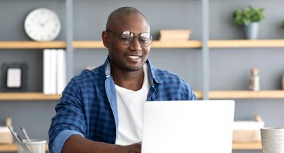 Smiling young man wearing round glasses and an open blue plaid dress shirt over a white tee works at his laptop in a home office