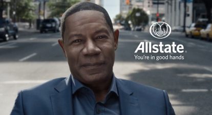 Actor Dennis Haysbert stands in the middle of a street looking into the camera next to the Allstate Insurance logo.