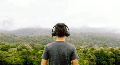 a man wth headphones on facing away from camera and looking out at a view of a lush green forest with low handing clouds.
