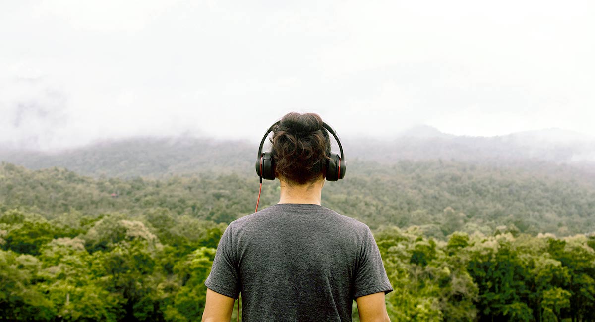a man wth headphones on facing away from camera and looking out at a view of a lush green forest with low handing clouds.