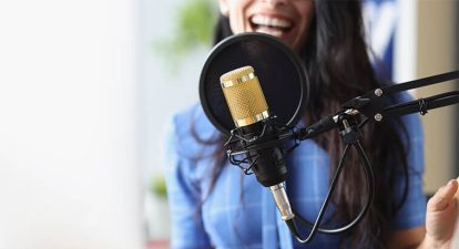 Woman in a blue shirt standing behing a gold mic and pop filter, delivering an audiobook narration read.