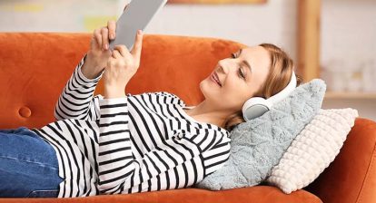 woman in a blakc and white striped shirt laying on an orange couch listening to an audiobook and looking at her cell phone.