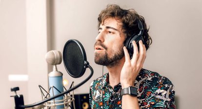 man in a colourful shirt standing at a mic with a pop filter, singing.