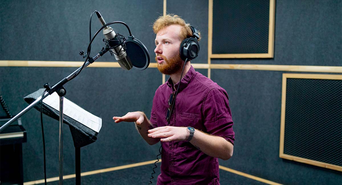 man with red hair and purple shirt in a voice over booth standing in front of a mic and pop filter.