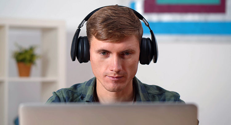 A white man with light brown hair wears headphones as he watches his computer in his home office.
