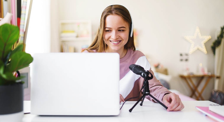 Smiling young woman sitting in front of a laptop and microphone recording a podcast.