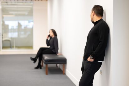 A woman listens to her phone on a bench inside, while a man looks towards her and leans against the wall in the hallway.
