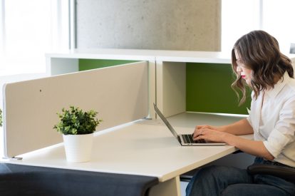 A white woman with brown hair works on her laptop on a white desk in an open concept office space.