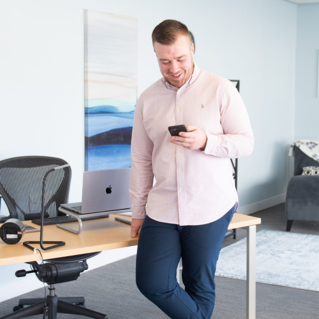 A white man wearing a pink shirt and jeans leans against his desk while checking his phone.