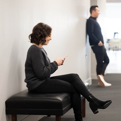 A woman with brown hair listens to her phone as she sits on a bench, a man leans against the wall further down the hall.