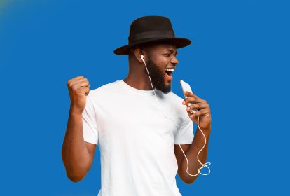 An african-american man wearing a black top hat sings into his phone in front of a blue background.