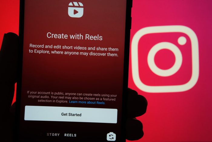 Instagram Reels start screen seen on mobile phone and Instagram logo on blurred background.