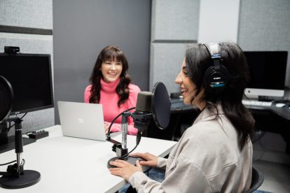 Two women wearing headphones and speaking into microphones in a studio.