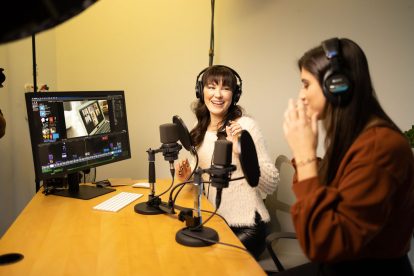 Two women wearing headphones smile and look at each other at they record a song in a studio.