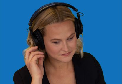 A blonde woman in front of a blue background listens to audio as she sits in front of a laptop.