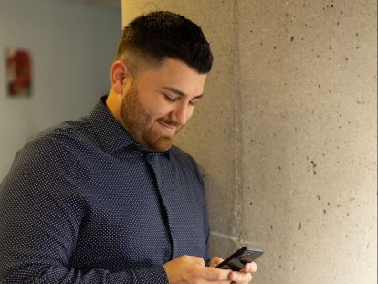 A man with dark hair leans against a pillar while looking at his phone.