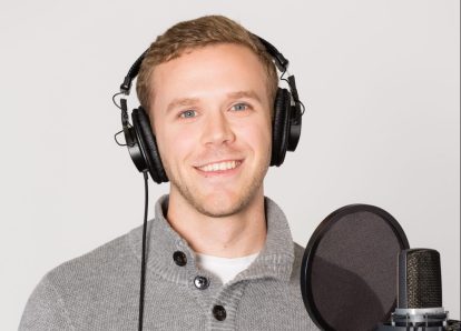 A blonde man wearing a grey shirt stands in front of a microphone with a pop filter.