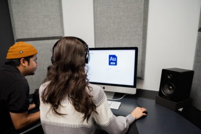 A man wearing an orange hat and a brunette woman sit in front of a computer editing audio files.