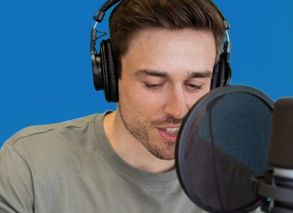 A man with brown hair looks at his phone and speaks into a microphone in front of a blue background.