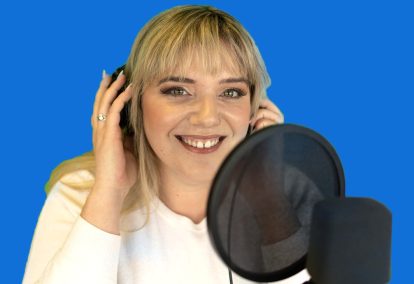 A blonde woman smiles while holding headphones to her ears and standing in front of a microphone.