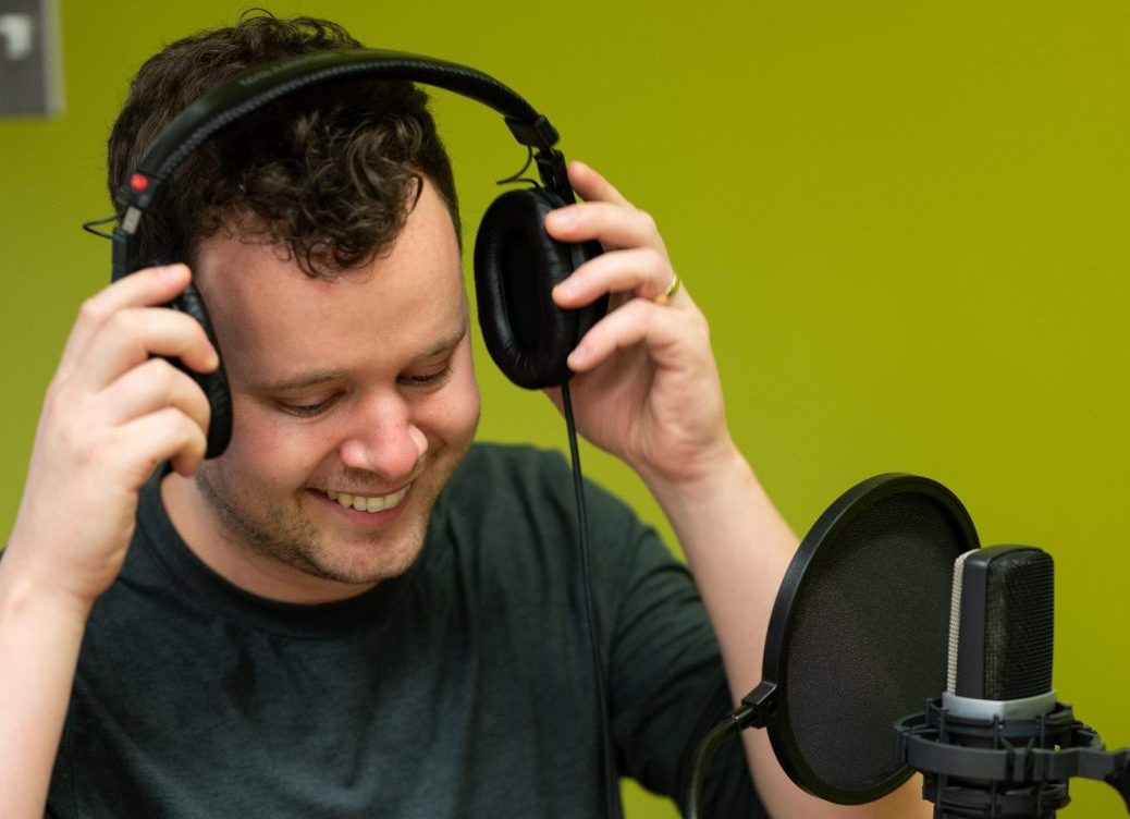 A man with black hair smiles towards a microphone as he puts on headphones.