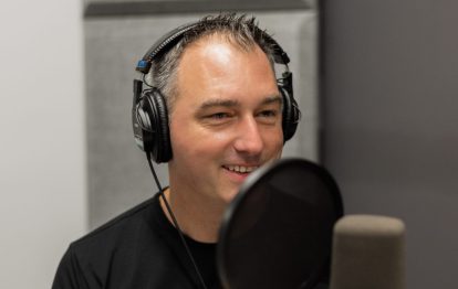 A man with grey hair and a black t-shirt smiles as he speaks into a microphone in a studio.