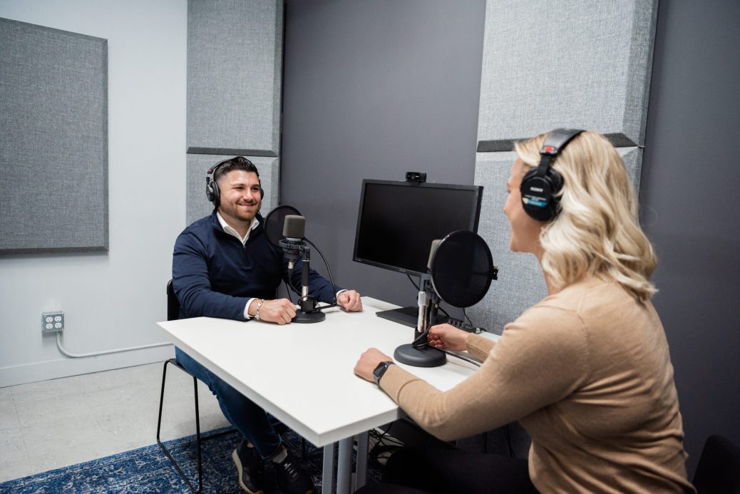 A man with dark hair and a beard smiles while wearing headphones and talking into a microphone while talking to a blonde woman doing the same.