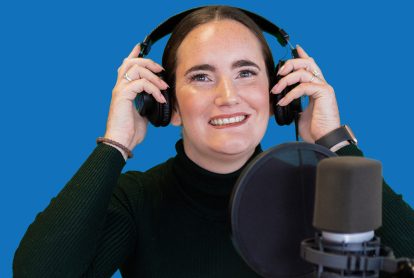 A brown haired woman smiles while wearing headphones and speaking into a microphone.