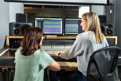 A blonde woman and brunette woman work near an audio editing board and computers in a recording studio.