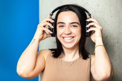 A woman with brunette hair holds black headphones to her ears as she smiles towards the camera.
