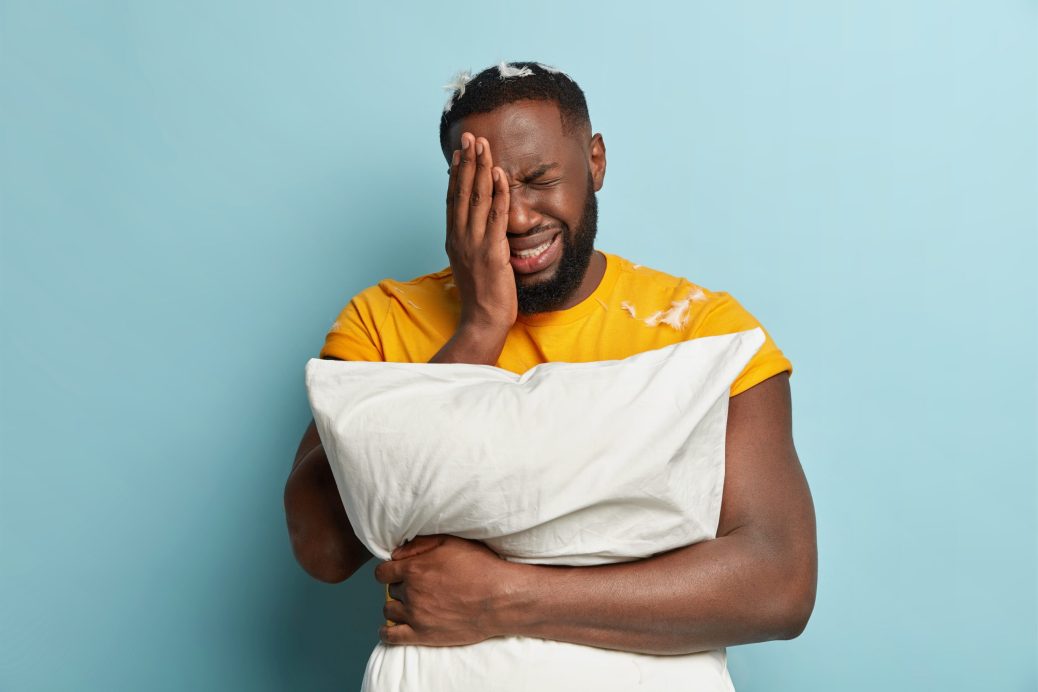 Dejected crying man covers face with hand, has thick stubble, dressed in yellow t shirt, has problems with sleep, carries white pillow, isolated over blue background.