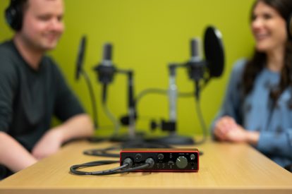 Two people smiling and speaking into a microphone with the camera in focus on the gain knob.