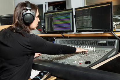 A picture of a woman in a black shirt wearing headphones working a studio board.