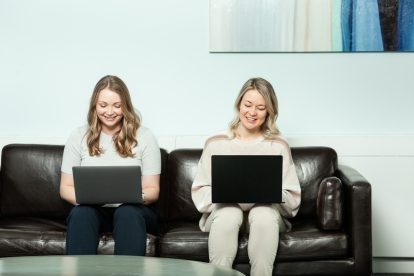 Two women sit on a couch working on their computers.