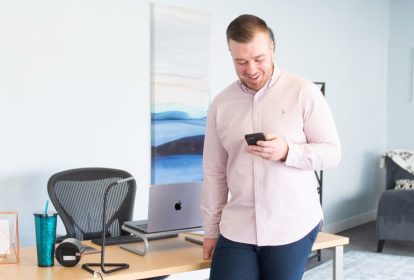 A man stands next to his desk looking at his phone.