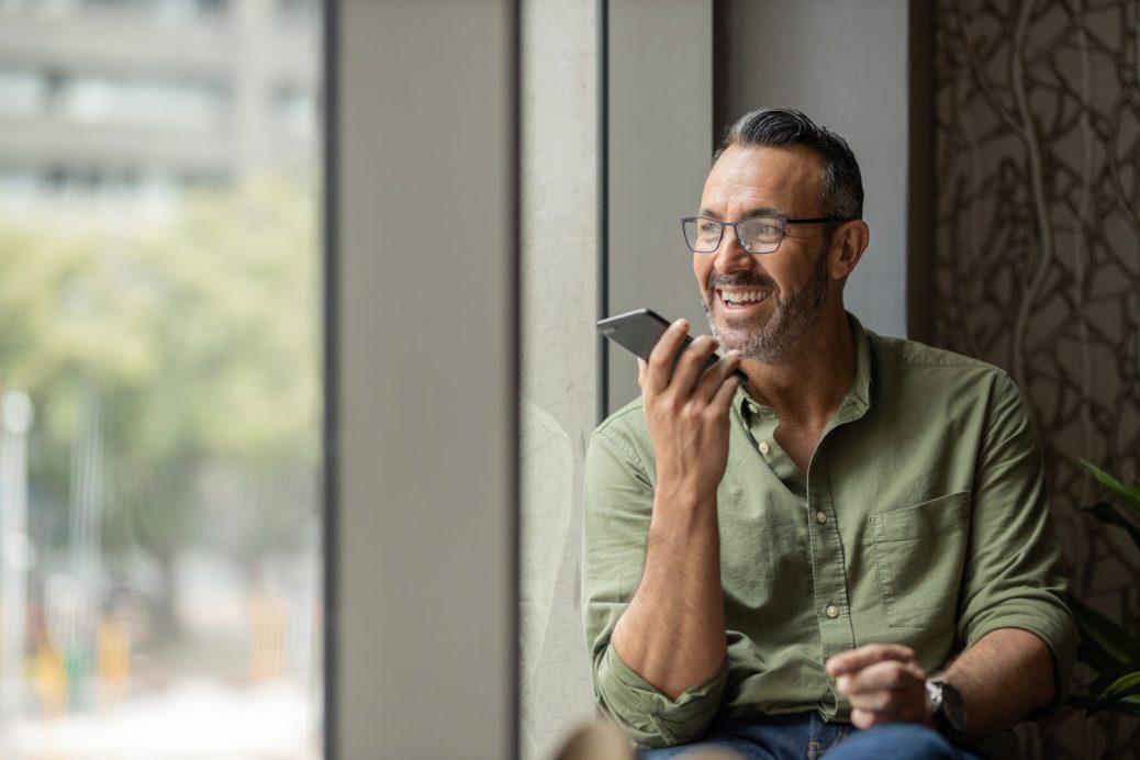 Wide angle image of Handsome mature bearded man sitting next to an looking out of sunny window talking on mobile smart phone smiling and wearing glasses