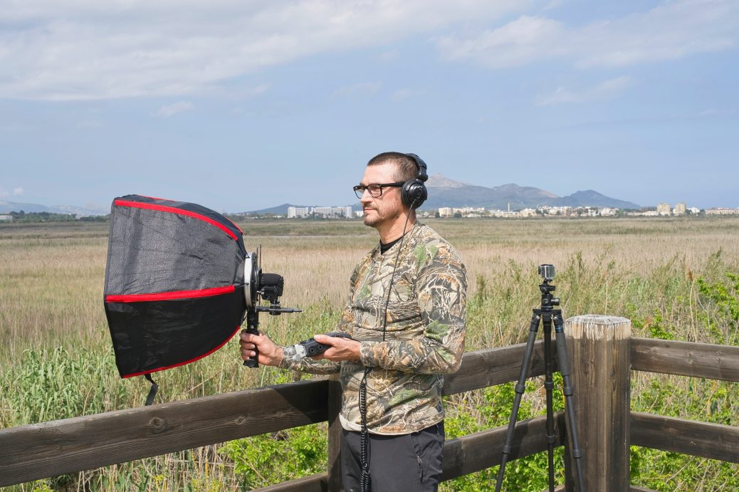 A man holds a parabolic microphone while standing outside beside a fence.