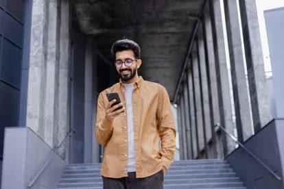 Successful young Hindu man walking outside office building, engineer software developer programmer smiling and happy using test app on phone, happy satisfied with result