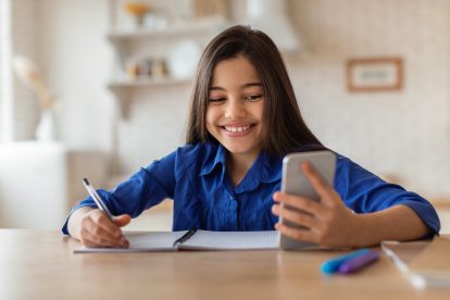 Cheerful Schoolgirl Using Cellphone And Taking Notes At Home