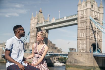 A young man and woman sitting on wall and smiling with London, England's Tower Bridge in the background.