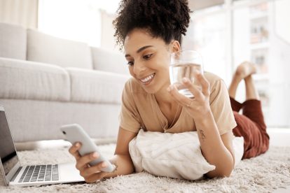 Relax, phone and black woman on social media for news reading or funny online content on living room floor. Smile, meme or happy girl enjoys a glass of water for wellness or healthy hydration at home.