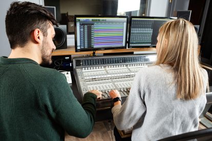A man with black hair and a woman with blonde hair sit beside each other working on an audio board in a studio.